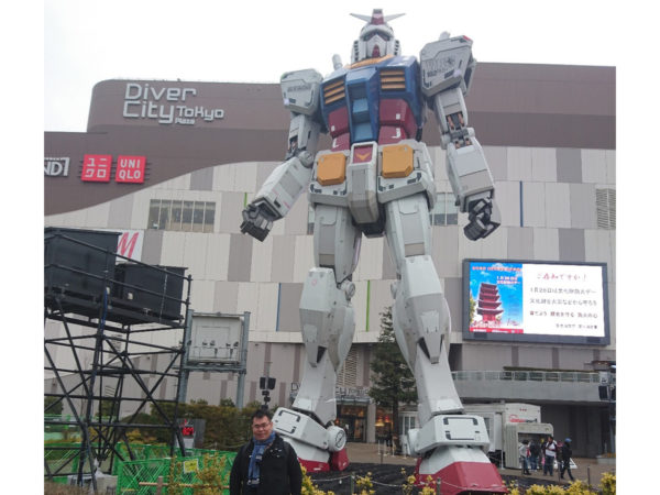 Desmond stojící před Diver City Tokyo Plaza s obří sochou Transformera za zády