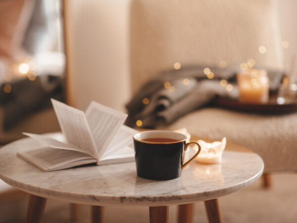 Kopje thee met papier open boek en brandende geurkaarsen op marmeren tafel boven gezellige stoel en gloeiende lichten in slaapkamer close-up. Winter vakantie seizoen.