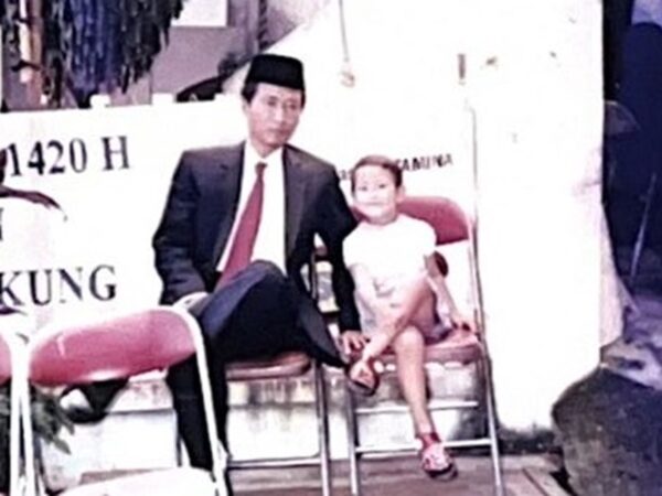 Mila, als kind, zittend op een stoel naast een man in een pak
