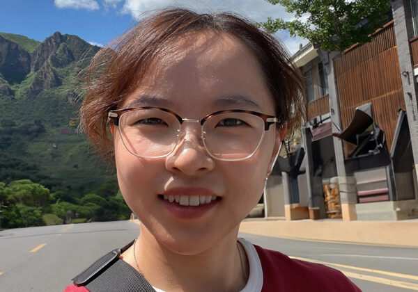 Η Yu Gao χαμογελάει στην κάμερα με ένα μεγάλο βουνό πίσω της