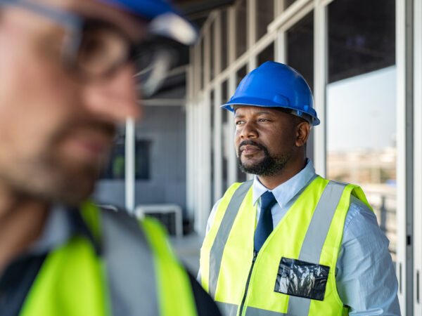 An African American man with a yellow vest and blue hard hat, looking out over a construction project