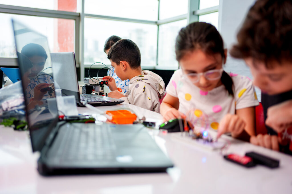 Group of young students participating in a hands-on robotics and STEM education activity. Two children are assembling a robot with various components, while others work on coding and programming using laptops. The classroom is vibrant and filled with educational tools and materials, promoting creativity, teamwork, and innovation.