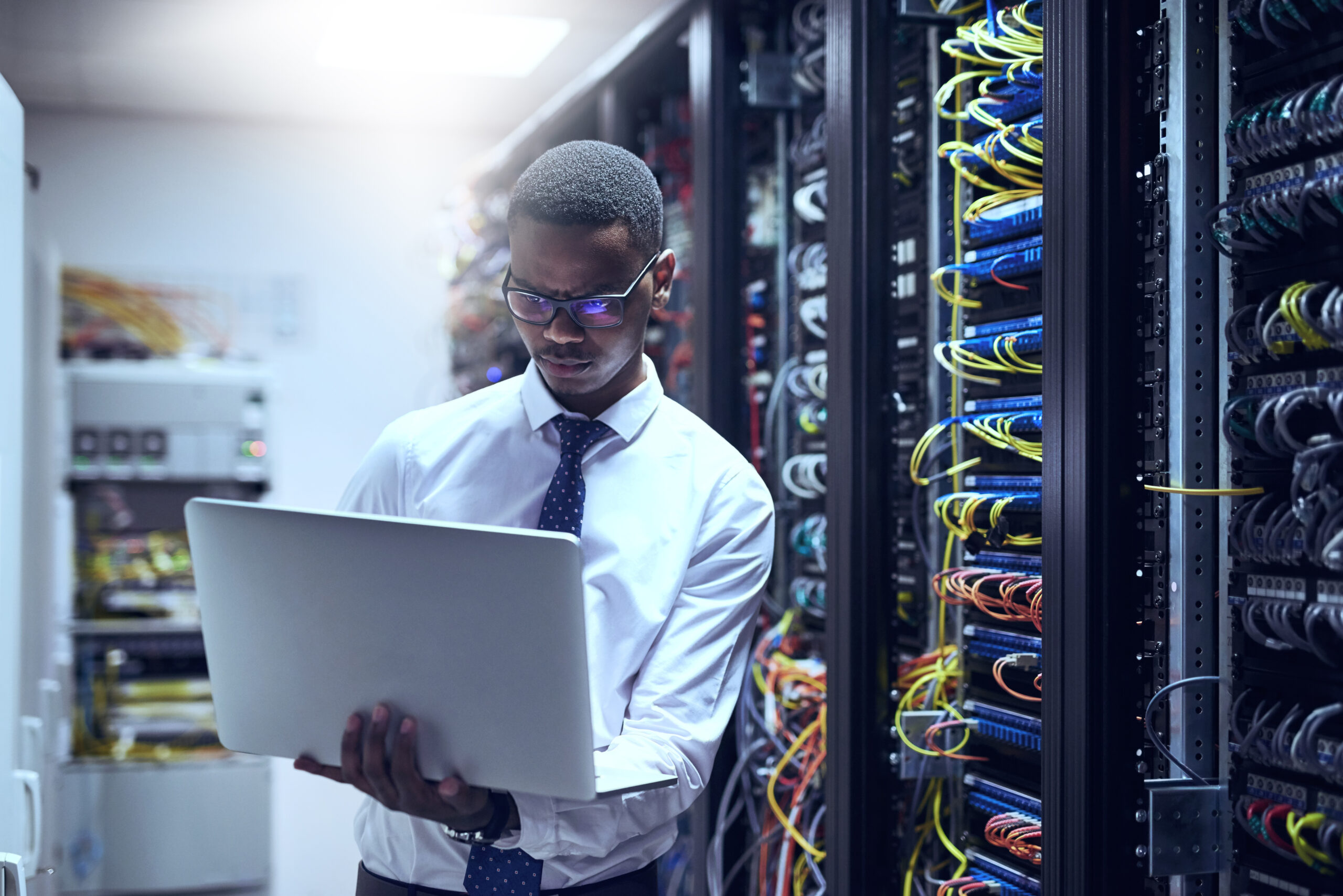 a young man standing in a server room working on a laptop