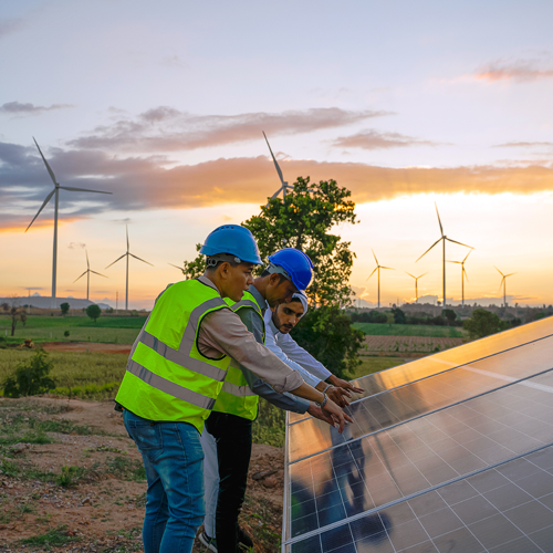 Two people working on solar panels with wind turbines in the distance at sunset