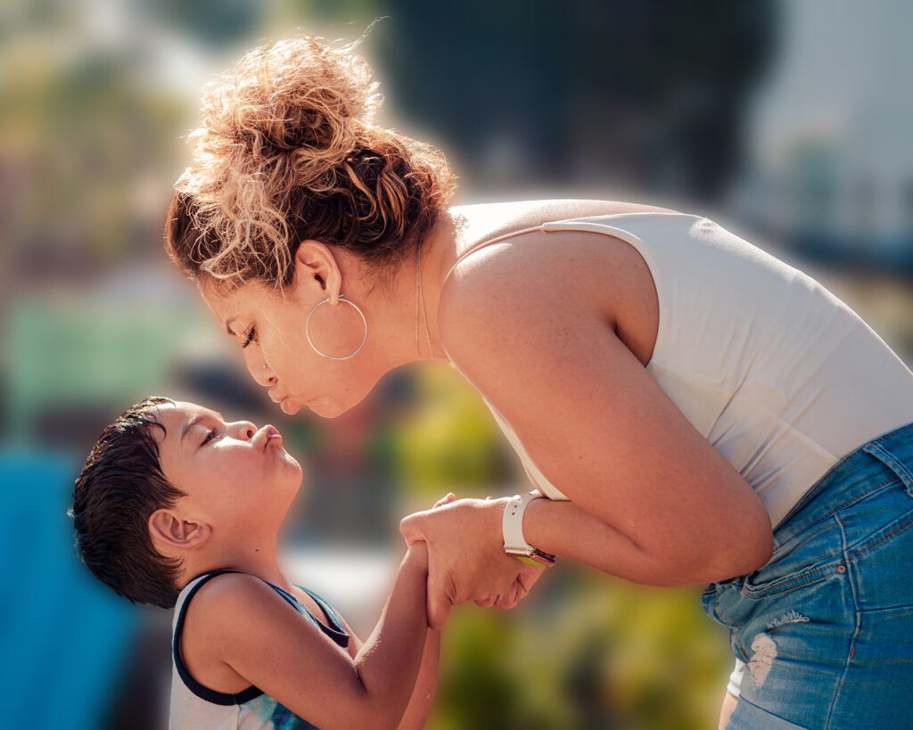 A Latina mother is leaning to kiss her son on the lips in an outdoor space